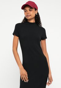 Sieviete ar cirtainiem matiem, tērpusies melnā ribotā īsroku kleitā un valkājot sarkankārtu beisbola cepuri, stāvot pret vienkāršu baltu fonu.