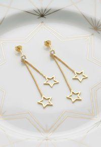 Boucles d'oreilles en or en forme de trois étoiles, ornées d'un cristal en haut. Des maillons de chaîne créent un mouvement, le tout présenté sur une plaque imprimée.