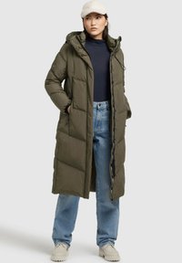 Lunga giacca a vento verde oliva con cappuccio, chiusura con zip e bottoni laterali. Abbinata a jeans blu e scarpe beige chiare.
