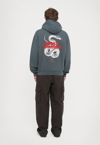 Haut à capuche gris foncé avec un graphique de serpent et le texte "SNAKE CHARMER" en rouge au dos. Associé à un pantalon cargo noir.