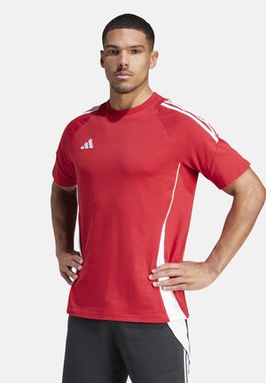 Uomo con maglietta sportiva rossa Adidas e pantaloncini neri in piedi con le mani sui fianchi davanti a uno sfondo neutro.