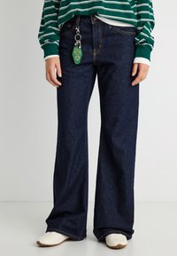 Άτομο που φοράει σκούρα μπλε τζιν με φαρδιά μπατζάκια, λευκά αθλητικά παπούτσια και πράσινο μακρυμάνικο πουκάμισο με ρίγες, με μπρελόκ να κρέμεται από το λουράκι της ζώνης.