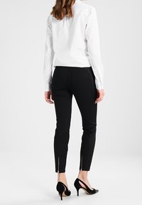 Vrouw staat, draagt een aansluitende zwarte broek met ritsen bij de enkels, een witte blouse en zwarte hoge hakken, met de rug naar ons toegekeerd.