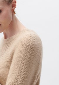 Maglione beige lavorato a maglia con motivi testurizzati, colletto a coste e dettagli in tessuto morbido e soffice sulle spalle. Orecchini dorati visibili.