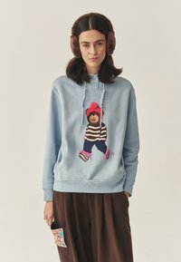 Világoskék kapucnis pulóver hímzett medve mintával, barna és rózsaszín részletekkel. Sötétbarna nadrággal és színes zseb részlettel párosítva.