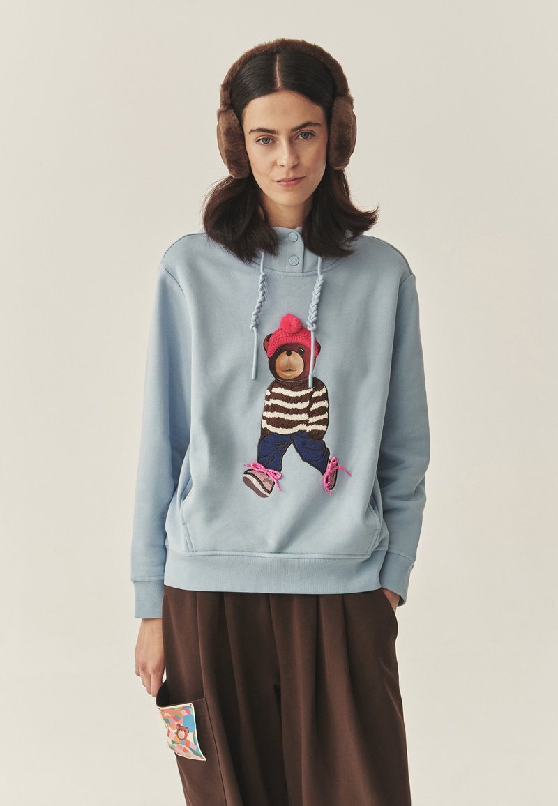 Világoskék kapucnis pulóver hímzett medve mintával, barna és rózsaszín részletekkel. Sötétbarna nadrággal és színes zseb részlettel párosítva.