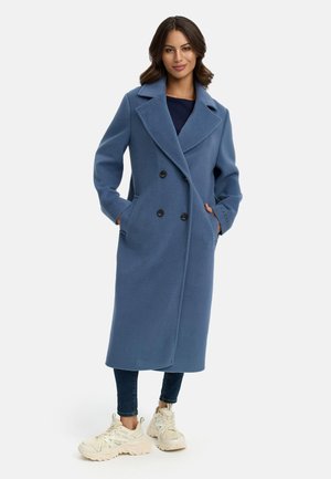 Cappotto lungo in misto lana blu con design doppiopetto, ampio colletto, due tasche frontali e bottoni neri. Abbinato a jeans aderenti e scarpe.