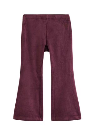 Ein junges Mädchen lächelt und steht mit gespreizten Beinen, trägt ein cremefarbenes, langärmeliges Oberteil mit Blumenmuster, lila Hose und braune Sneakers mit roten Schnürsenkeln.