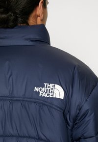 Person trägt eine marineblaue Steppjacke mit dem Logo "The North Face" auf dem oberen Rücken, von hinten sichtbar.