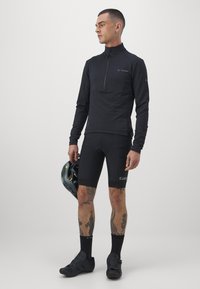 Musthave musta pika varrukaga jalgrattasärk poole tõmbega, koos mustade jalgrattapükstega. Sile kangas, kitsas lõige ja sportlik disain. Kiiver käes.