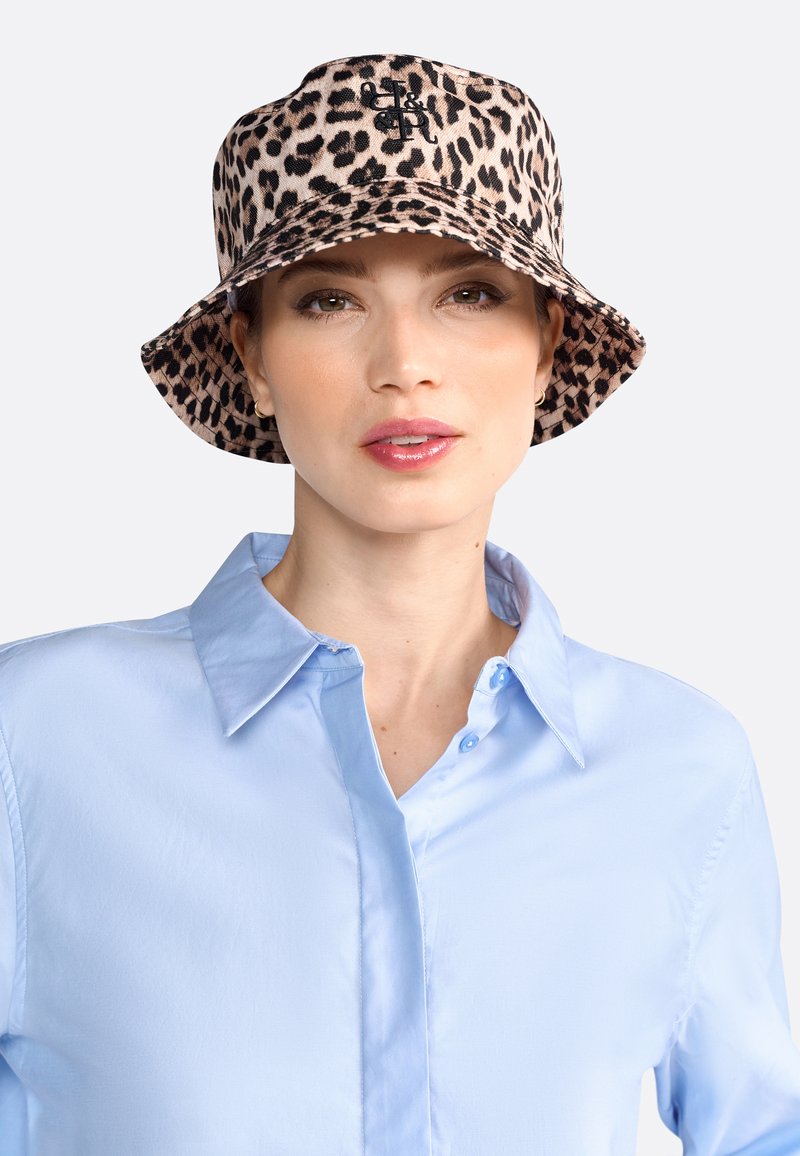 Beige Bucket Hat aus Stoff mit schwarzem Leopardenmuster; besitzt ein Logo auf der Vorderseite. Kombiniert mit einem hellblauen Hemd mit Kragen.