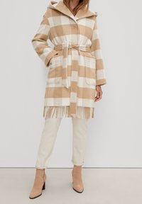 Žena v bežovém a bílém kostkovaném kabátu s páskem a třásněmi, krémových kalhotách a světlých semišových kotníčkových botách stojí.