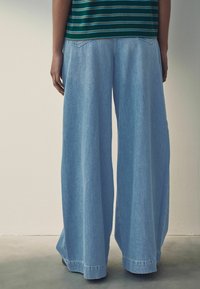 Pantalones de mezclilla de pierna ancha en azul claro con dobladillo sin rematar y bolsillos traseros, combinados con una camisa de rayas verde. Textura suave y ajuste holgado.