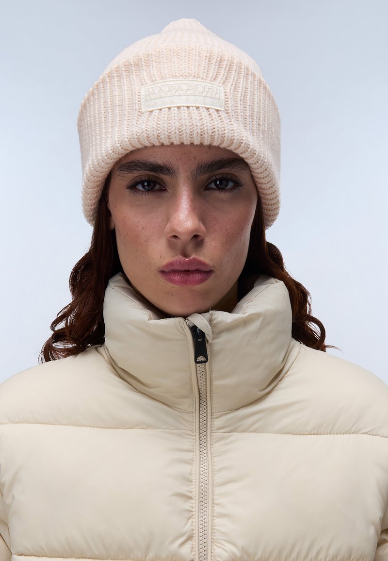 Berretto lavorato a maglia beige chiaro con texture a costine e applicazione di logo, abbinato a un giubbotto imbottito beige con colletto alto e zip.