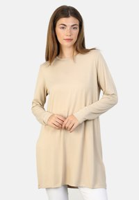 Tunique à manches longues beige avec un col rond, fabriquée en tissu doux, présentant une coupe décontractée et un ourlet légèrement arrondi.