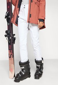 Λευκό σκι παντελόνι με λεία υφή, διαθέτει ψηλή μέση και κλείσιμο με δύο κουμπιά, συνοδευόμενο από μαύρες μπότες σκι και ένα σκι.