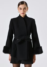 Cappotto di lana nera con design avvolgente, vita cintrata e dettagli in pelliccia sintetica sui polsini. Presenta linee pulite e una finitura testurizzata.