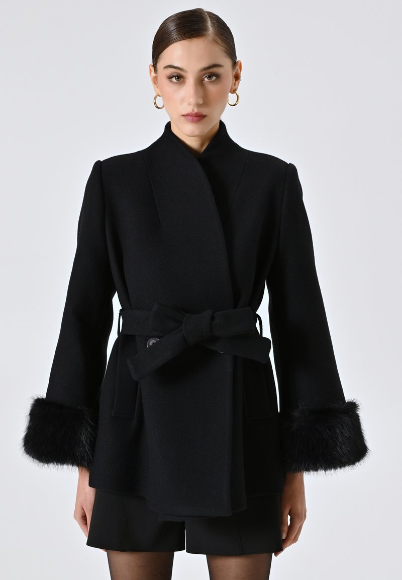 Cappotto di lana nera con design avvolgente, vita cintrata e dettagli in pelliccia sintetica sui polsini. Presenta linee pulite e una finitura testurizzata.