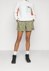 Sudadera de felpa blanca con acentos en azul claro y coral, combinada con shorts con estampado en verde y naranja. Botas de senderismo negras y calcetines blancos.