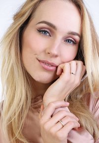 Gouden ring met kleine edelsteen, gedragen aan de rechterhand, terwijl het model poseert met de hand bij het gezicht. Glad huid, lang haar en zachte verlichting.