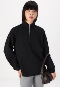 Svart quarter-zip genser med overdimensjonerte ermer og en avslappet passform, båret over lysegrå leggins. En liten brun veske holdes i hånden.