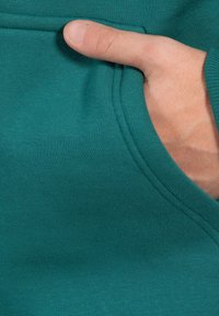 Sudadera de color azul verdoso con un detalle de bolsillo. Textura suave con costuras visibles y una mano parcialmente dentro del bolsillo, mostrando su funcionalidad.