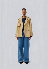 Γυναίκα που στέκεται με το πρόσωπο μπροστά, φορώντας ένα μπεζ διπλόσακο, μαύρη μπλούζα και φαρδιά γαλάζια τζιν παντελόνια με μαύρες γόβες.