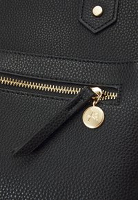 Černá kožená taška s texturovaným povrchem, zlatým zipem a kulatým kovovým přívěskem s vyraženým logem.