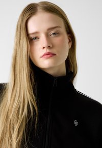 Czarna kurtka zapinana na zamek z wysokim kołnierzem, srebrnym logo na piersi i kontrastowymi paskami na ramionach. Gładka faktura tkaniny.