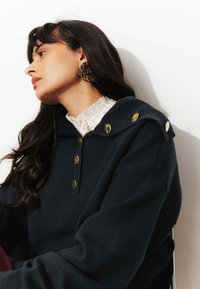 Sweatshirt navy avec capuche, détails en boutons dorés et col texturé. Le modèle porte de grandes boucles d'oreilles à motifs. Texture de tissu lisse.