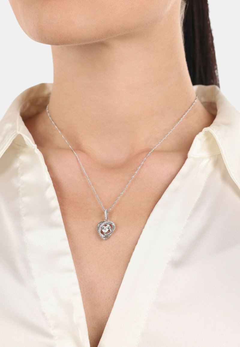Silverfärgad hjärtformad hängsmycke med en kristall i mitten och prydd med små stenar på en smäcker kedja, bärs över en ljus blus.