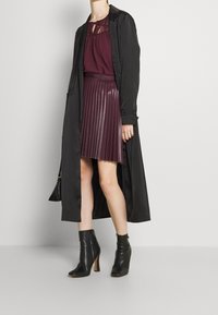 Sort lang frakke over en bordeaux top med blonder og en plisseret bordeaux skørt i kunstlæder. Sorte ankelstøvler fuldender outfittet.