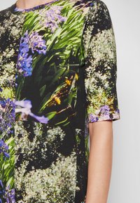 Gros plan d'une chemise à manches courtes avec un imprimé floral coloré comprenant des feuilles vertes, des fleurs violettes et des fleurs blanches.