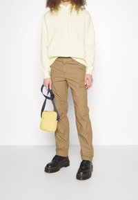 Person som bär en ljusgul Nike-hoodie, beige cargobyxor, svarta platåskor och håller en liten gul crossbody-väska med ett marinblått band.