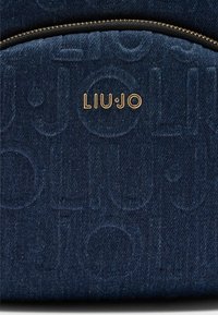 Marineblauwe denim rugzak heeft een opvallend reliëflogo, gouden hardware-accenten en een ritssluiting. Zachte textuur.
