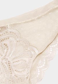 Beige kanten lingerie met bloemmotieven en gegolfde randen op doorschijnende mesh stof.