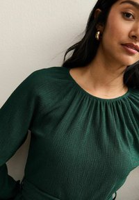 Zielona bluzka o fakturowanej powierzchni z okrągłym dekoltem i marszczonymi detalami na ramionach. Długie rękawy, w zestawie z pasującym paskiem.