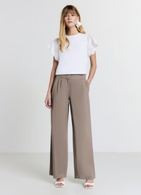 Chemise blanche avec manches en dentelle ajourée, associée à un pantalon taupe taille haute à jambes larges. Le mannequin porte des sandales blanches à talons.