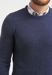 Mörkblå stickad tröja med rund halsringning. Tyget har en strukturerad yta, synlig ovanför en mönstrad vit skjortkrage.