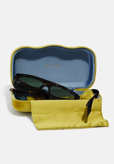 Gafas de sol Gucci en un marco de carey oscuro, con lentes verdes, dentro de un estuche blando azul con ribete amarillo, acompañado de una bolsa de satén amarilla.