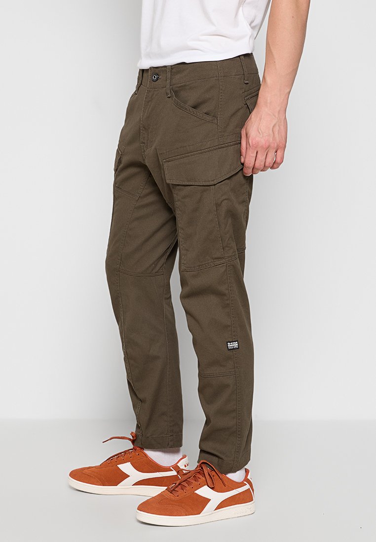 Pantalones cargo verde oliva con múltiples bolsillos, tejido resistente, corte recto y una etiqueta de marca sutil en el muslo. Combinados con zapatillas naranjas.