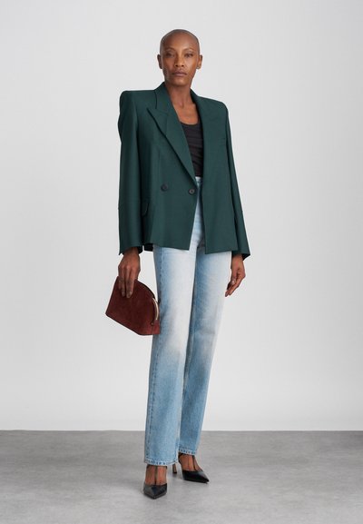 Σκοτεινό πράσινο σακάκι με διπλό κούμπωμα και δομημένα ώμους, σε συνδυασμό με ανοιχτό μπλε τζιν σε ευθεία γραμμή και μαύρες γόβες με μυτερή μύτη, κρατώντας μια μπορντό τσάντα.
