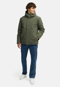 Olivengrøn isoleret jakke med lynlås og trykknaplukning, hætte, to frontlommer og en afslappet pasform, parret med blå jeans og hvide sko.