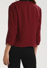 Burgundy jacket with three-quarter sleeves, smooth texture, and a back seam detail; worn over black pants.
