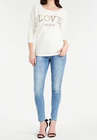 Top bianco a maniche lunghe con le scritte "LOVE" e "GUESS" decorate con paillette, abbinato a jeans skinny blu chiaro e sandali neri con tacco.