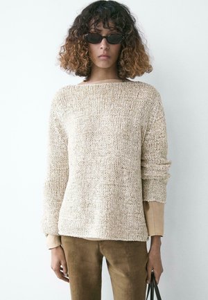 Personne aux cheveux bouclés mi-longs portant des lunettes de soleil ovales noires, un pull en grosse maille beige, un pantalon marron clair et tenant un sac noir.