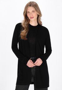 Cardigan lungo nero realizzato in tessuto morbido, con design aperto davanti, maniche lunghe e una vestibilità rilassata. Indossato sopra una maglietta nera e jeans.
