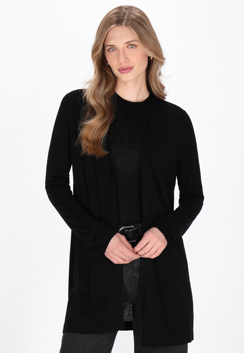 Cardigan lungo nero realizzato in tessuto morbido, con design aperto davanti, maniche lunghe e una vestibilità rilassata. Indossato sopra una maglietta nera e jeans.