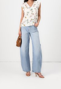 Nainen, jolla on päällä valkoinen kukallinen hihaton toppi, vaaleansiniset leveälahkeiset farkut, vaaleat korkokengät ja käsissään ruskea käsilaukku.