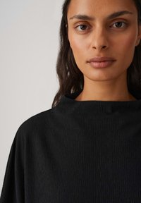 Schwarzes, strukturiertes Kleidungsstück mit hohem Kragen und einem gerippten Stoffmuster. Das Design zeichnet sich durch breite Ärmel aus und zeigt eine lässige Silhouette.